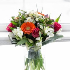 This floral composition, reminiscent of the sophisticated greenery and community feel of Denmark Hill, is a striking study in colour, form, and local charm. Centred within a tall glass vase, creamy peach and vibrant orange gerberas radiate outward, their soft petals and rich brown centers glowing in gentle daylight. Deep magenta roses, petals wound tight, introduce layers of opulence and heartfelt romance. White alstroemeria and petite spray roses thread among the larger blooms, adding lightness and the freshness so often found in local community gardens. Pastel pink and pale filler flowers provide soft highlights and nuances, their placement careful and artistic. Broad, glossy aspidistra leaves form a sculptural collar at the bouquet's base, echoing the well-tended parks and green spaces that define Denmark Hill. Interlacing smaller leaves add depth and fullness, while the clear vase reveals an array of sturdy green stems. The natural light enhances each colour and texture, lending the entire display a sense of renewal and celebration-a perfect gift for welcoming neighbours, marking anniversaries, or adorning a Denmark Hill home with local flair and expert touch.